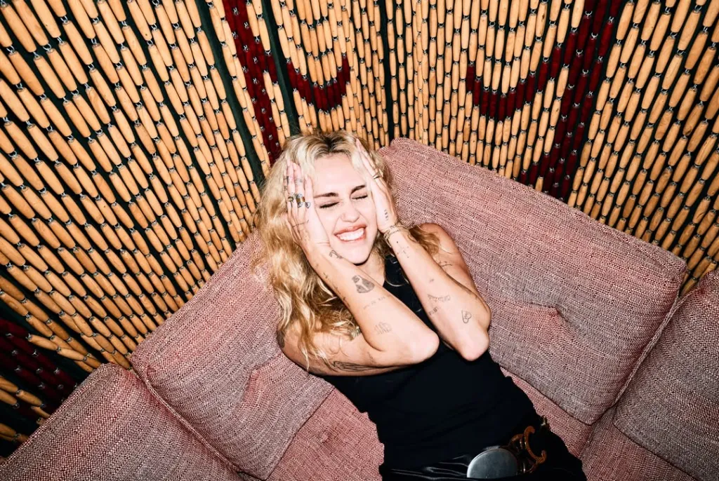 Miley Cyrus sits on a pink couch in front of a wooden bead curtain, holding her head in her hands and smiling.