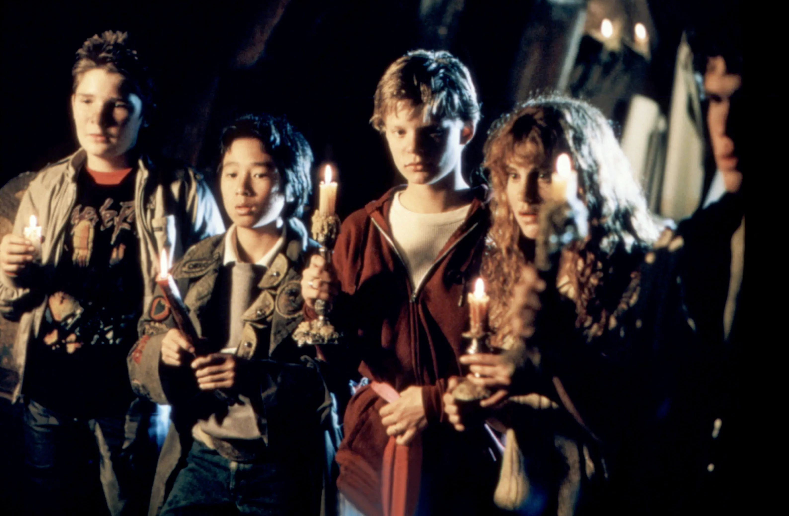 A scene from The Goonies with Corey Feldman, Ke Huy Quan, Martha Plimpton, Kerri Green, and Josh Brolin holding candles in the dark.