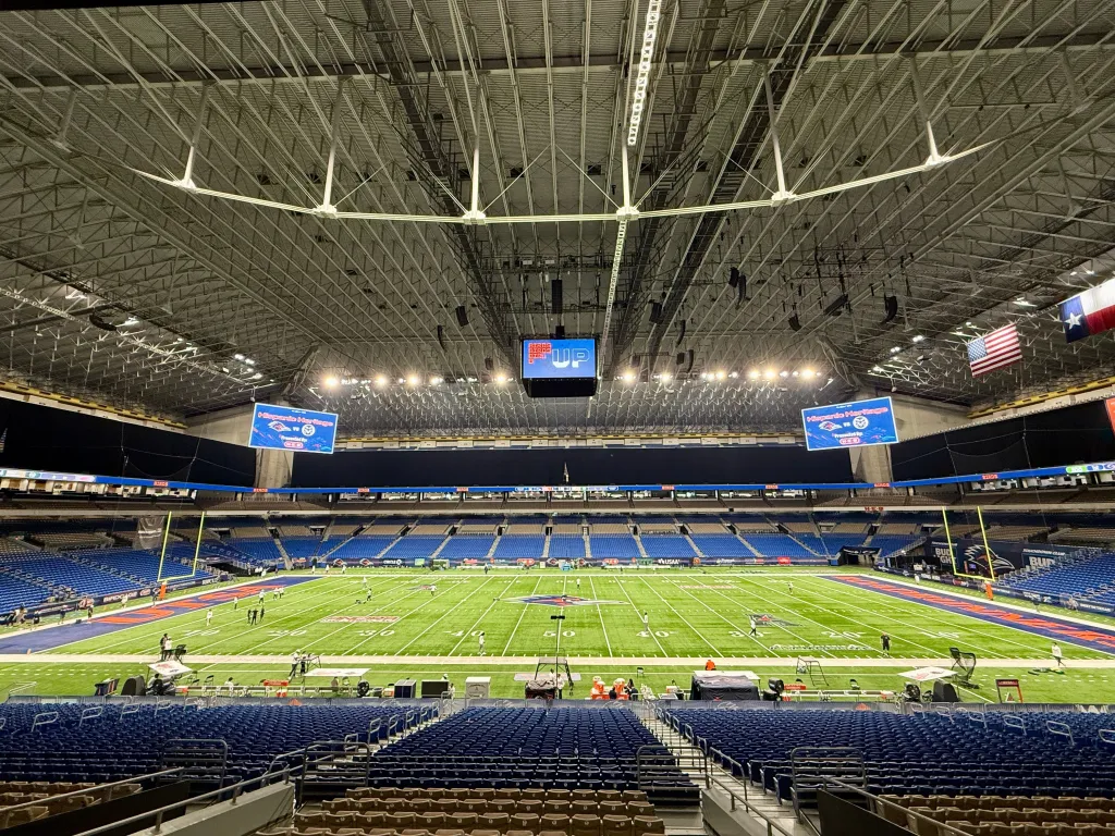 An interior view of the Alamodome showing the football field and seating rows under the stadium's high arched roof.