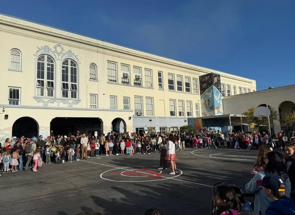 A large crowd of children and adults gathers in the asphalt courtyard of Alvarado Elementary School in San Francisco.