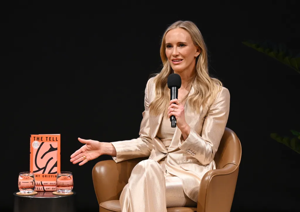 Amy Griffin speaks onstage holding a microphone while seated in a chair next to a table with a copy of her book, 