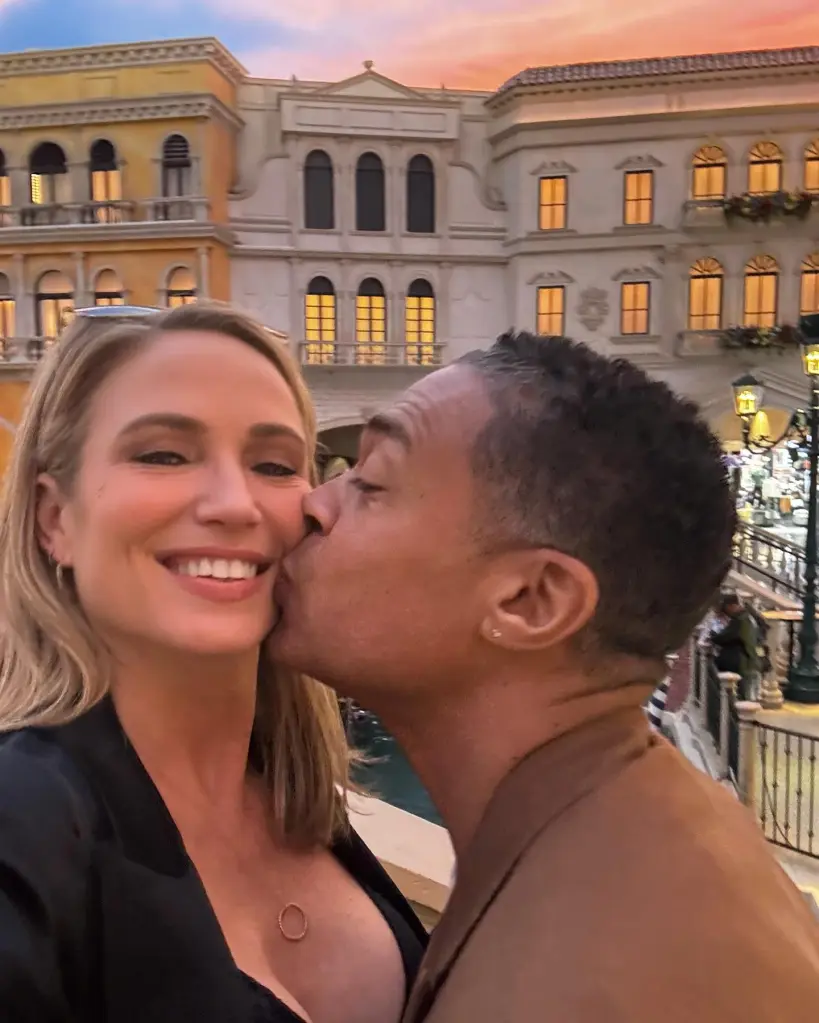 Amy Robach smiling as T.J. Holmes kisses her cheek in front of European-style buildings.