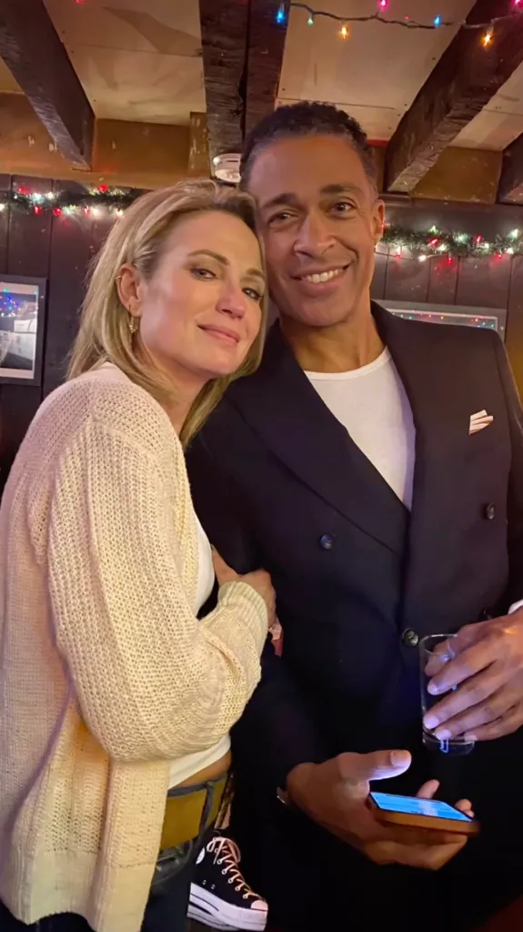 Amy Robach and T.J. Holmes smiling together indoors with festive lights in the background.