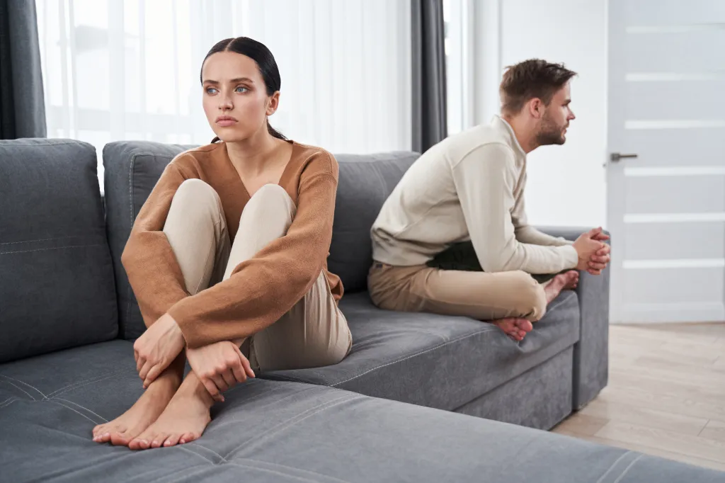 A young couple sits silently at opposite ends of a sofa