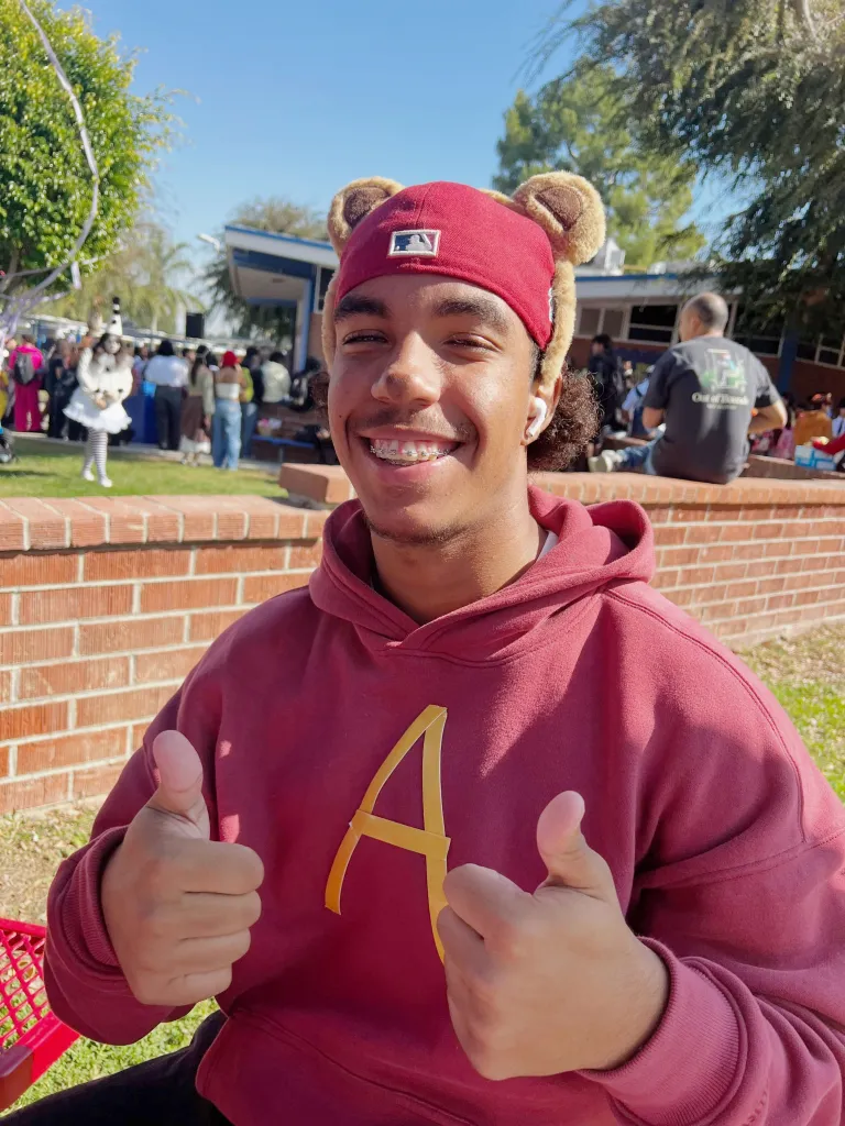 Anthony Joseph Blas giving a double thumbs-up while wearing a maroon hoodie with a yellow 'A' and a red hat with bear ears outdoors.