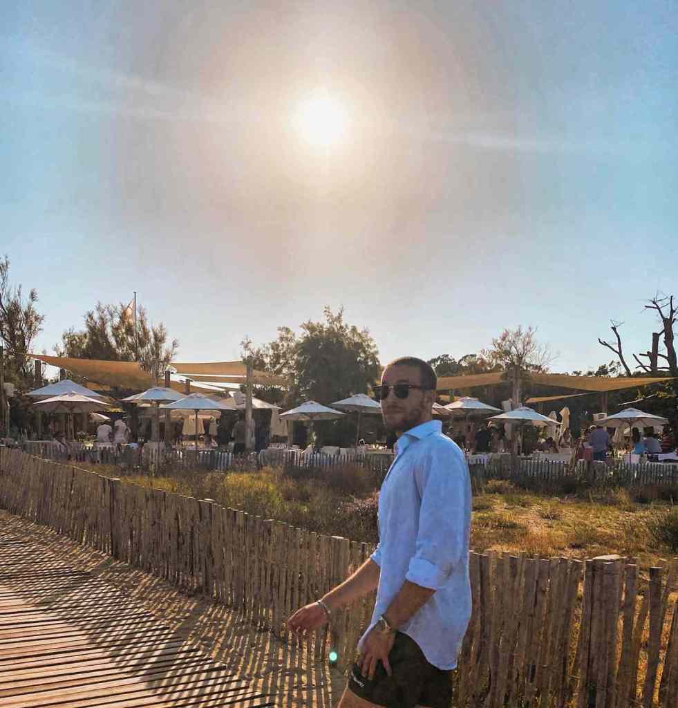 Antoine de Massy walking on a wooden boardwalk by the beach in the afternoon sun.