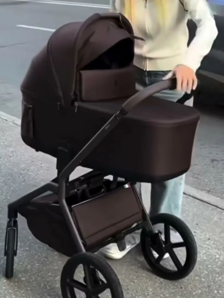 A person pushing a brown baby stroller containing a portable speaker on a sidewalk.