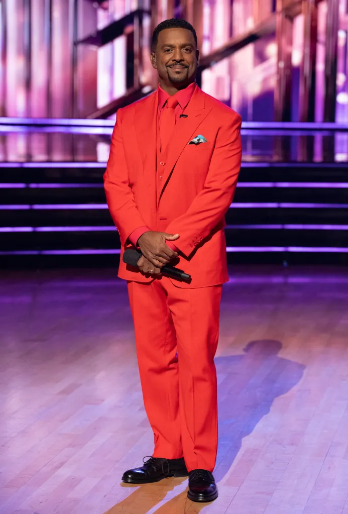 Alfonso Ribeiro wearing a red suit while holding a microphone on the Dancing with the Stars ballroom floor.