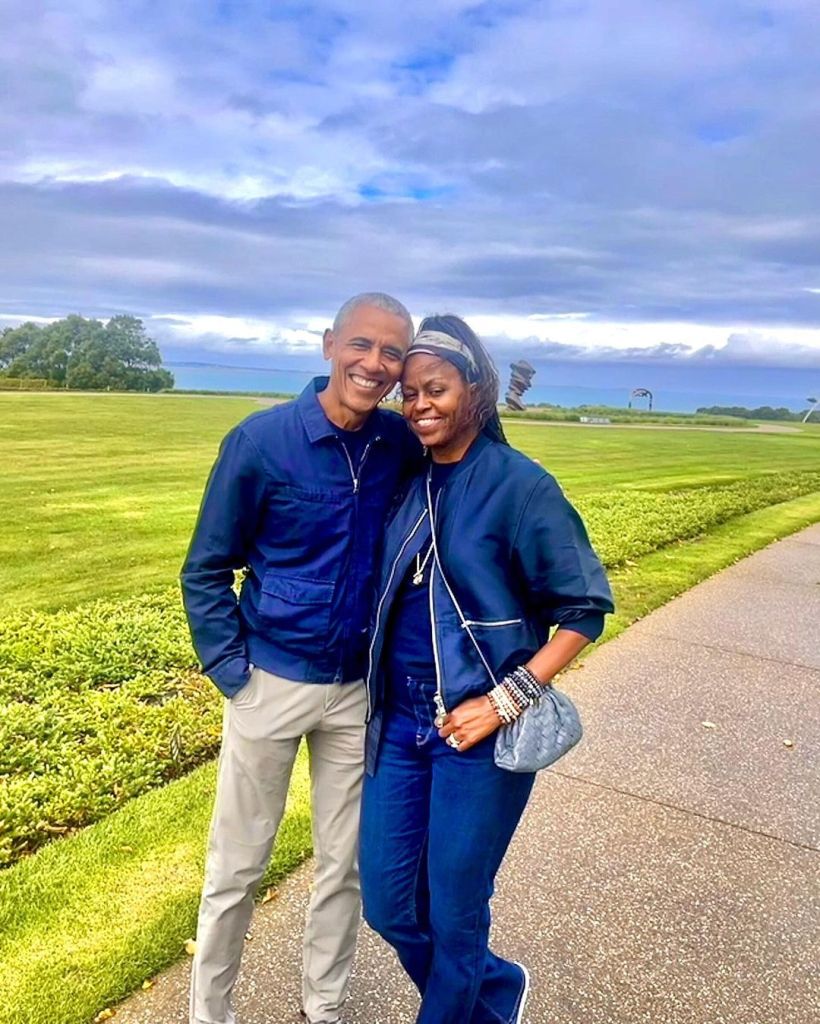 Barack and Michelle Obama stand close together outdoors, smiling and embracing, with a grassy landscape and cloudy sky in the background.