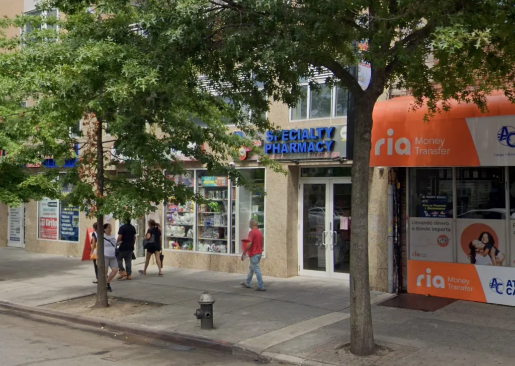 Exterior view of Baron Specialty Pharmacy in Jackson Heights, N.Y., with pedestrians walking on the sidewalk.