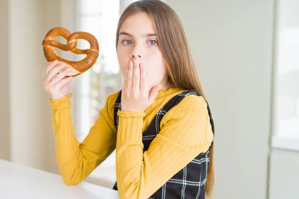 A young girl holding a pretzel looks shocked, covering her mouth with her hand.