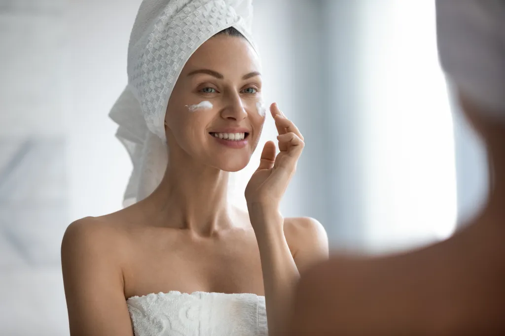 A young woman with a towel on her head smiles as she applies facial lotion in a mirror.