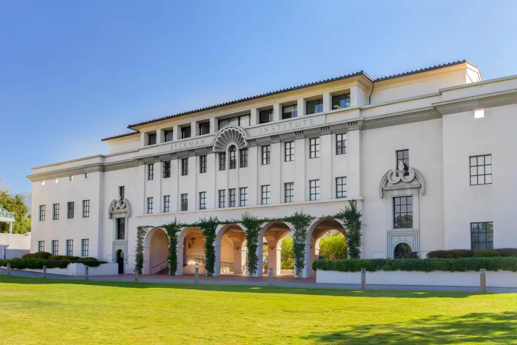 The Beckman Institute building on the California Institute of Technology campus, featuring a pale exterior, arched ground-floor walkway with climbing vines, and a large lawn in the foreground under a clear blue sky.
