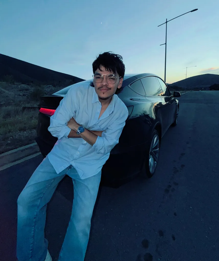 Twitch streamer Komanche posing with his arms crossed while leaning against a black car parked on a road at dusk.