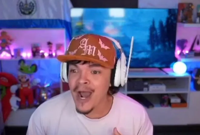 Twitch streamer Komanche wearing a baseball cap and white headphones while streaming in his room.