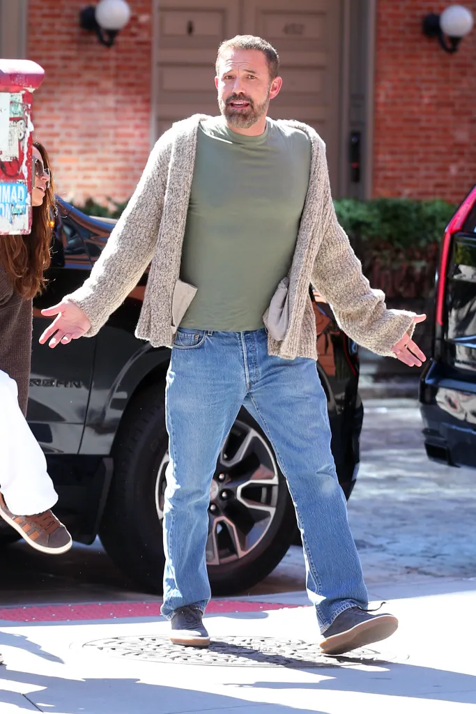 Ben Affleck walking down a New York City street in a green t-shirt, grey vest, and blue jeans, with Luciana Barroso partially visible next to him.