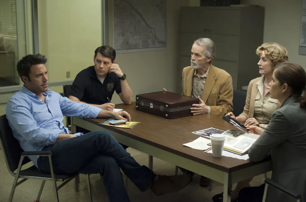Ben Affleck, Patrick Fugit, David Clennon, Lisa Banes, and Kim Dickens seated around a table in a police station interrogation room in a scene from the film Gone Girl.