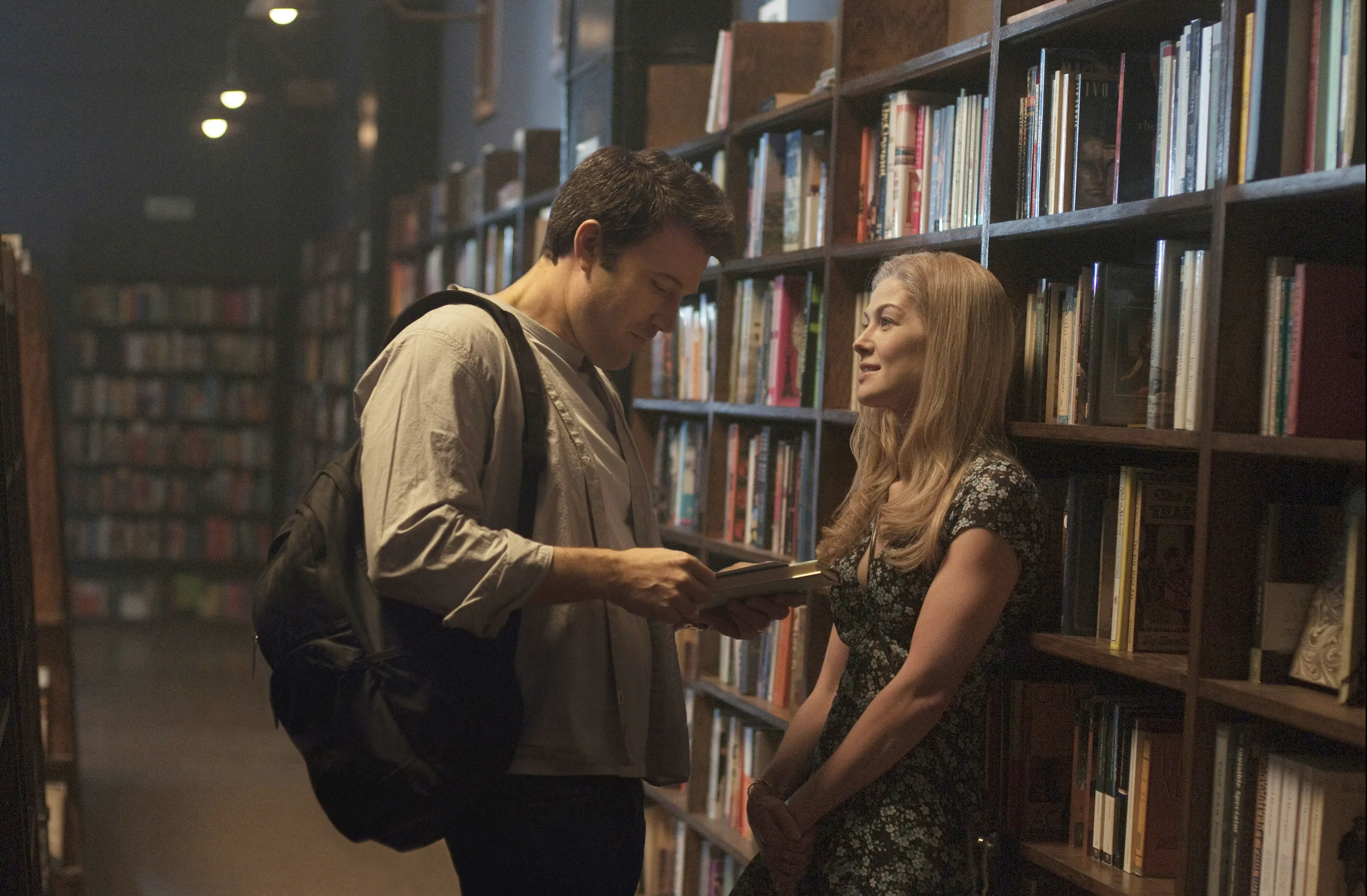 Ben Affleck and Rosamund Pike standing in a bookstore aisle, with Ben holding a book and looking at Rosamund.