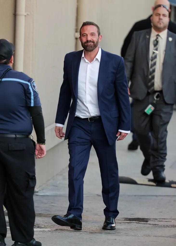 Ben Affleck wearing a dark blue suit and white shirt, flanked by security personnel.