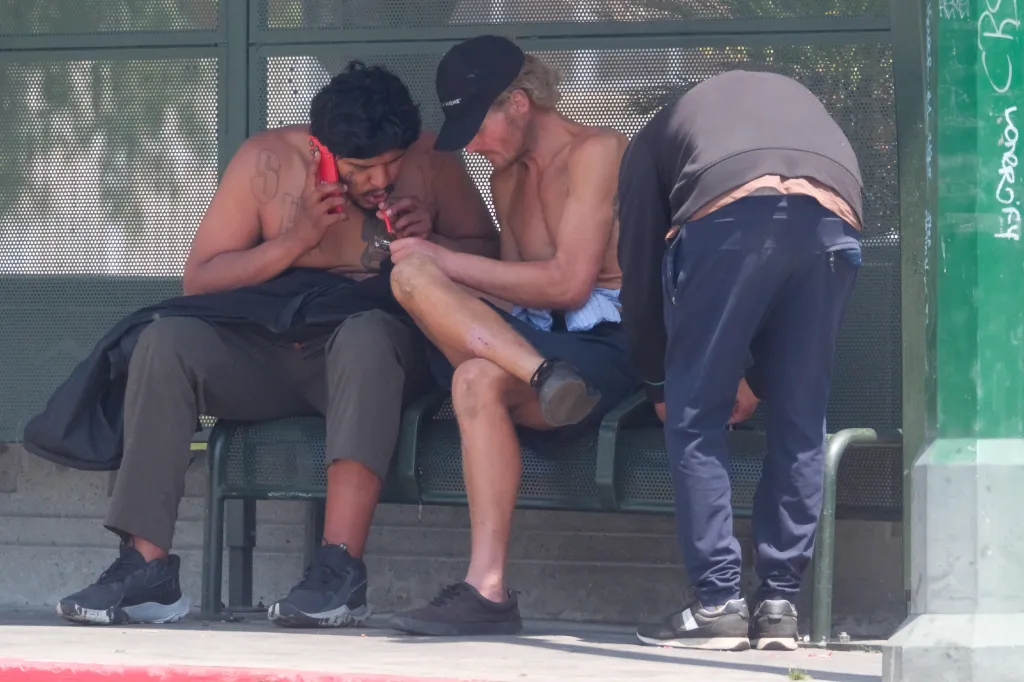 Three men gather on and around a green bus stop bench, with one smoking from a pipe.