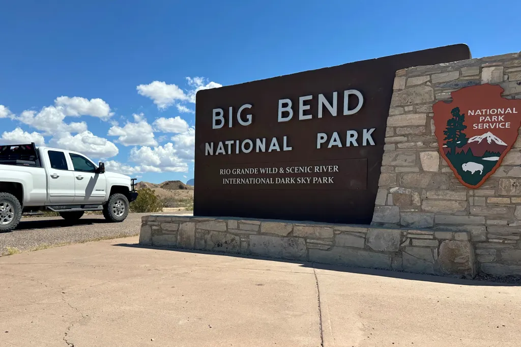 Entrance sign for Big Bend National Park in Texas, with a white pickup truck parked nearby under a blue sky.