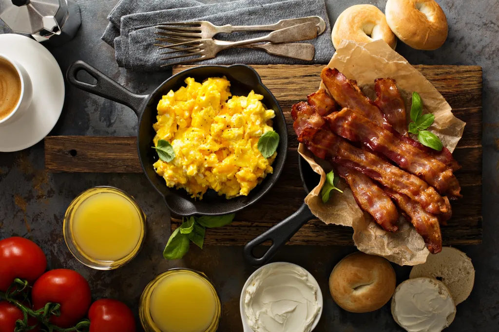 Overhead view of a hearty breakfast with scrambled eggs in a skillet, crispy bacon, bagels, orange juice, and coffee.