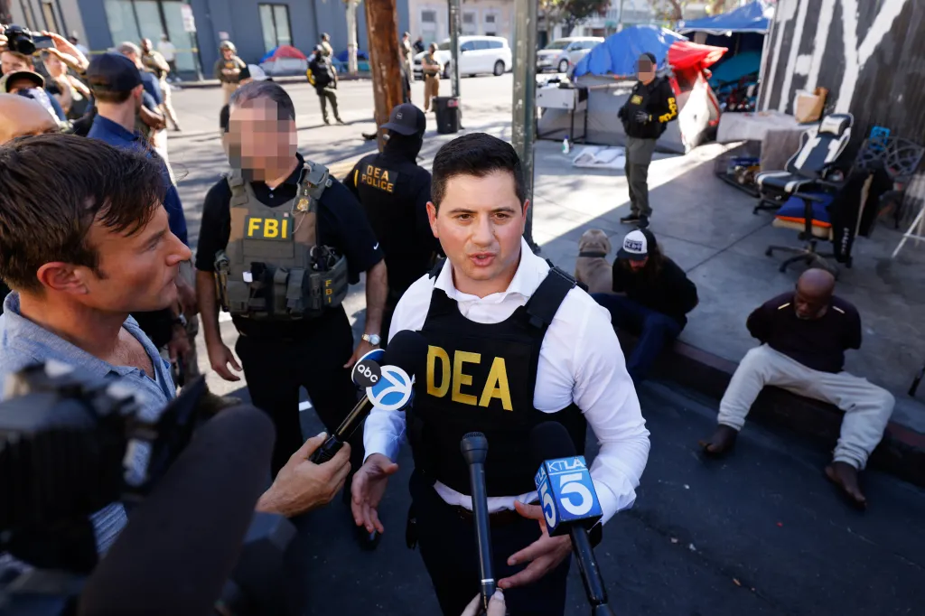 Bill Essayli speaks to reporters during Operation Free Skid Row, flanked by law enforcement officers with detained individuals seated in the background.