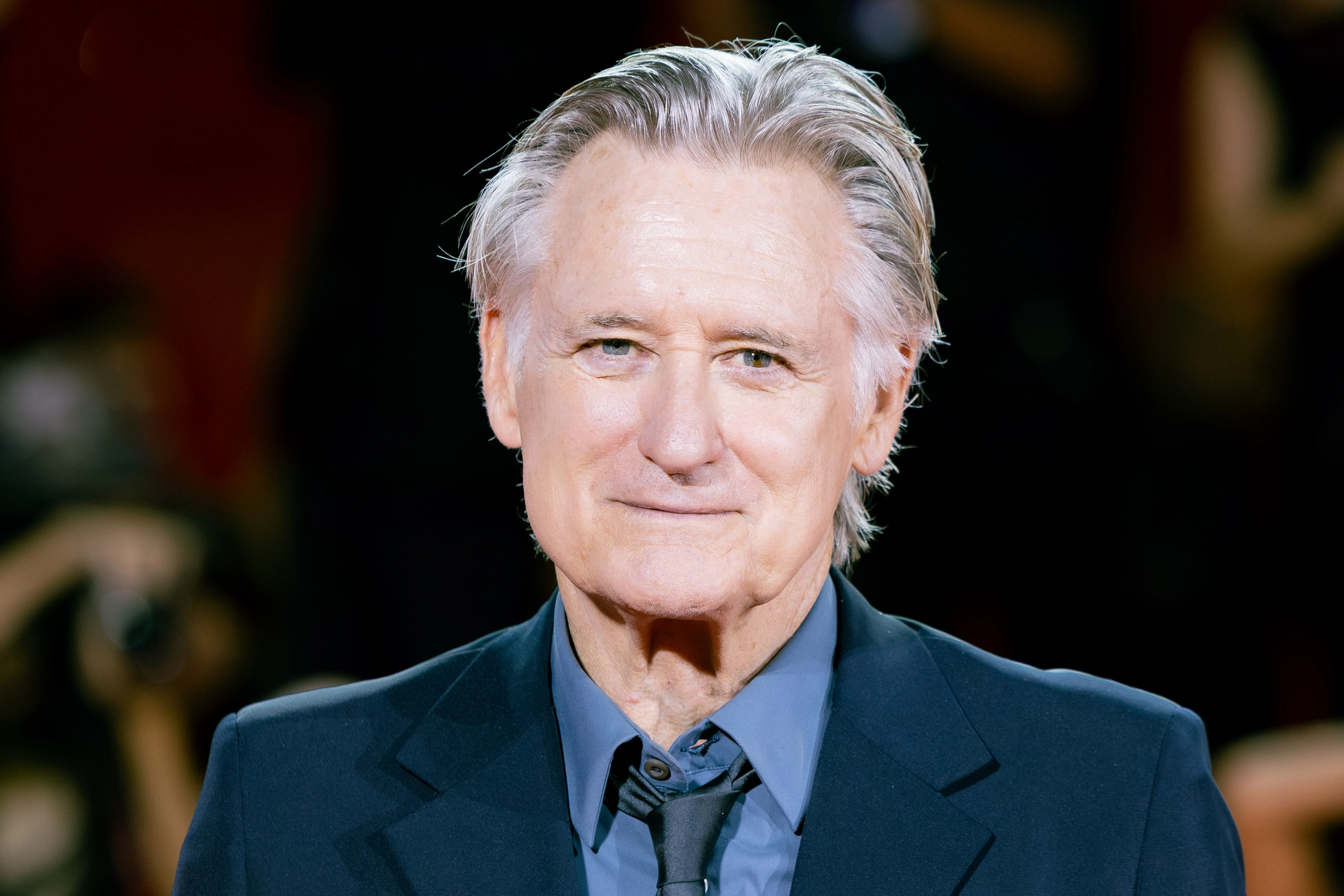 Actor Bill Pullman smiling in a dark suit and blue shirt.