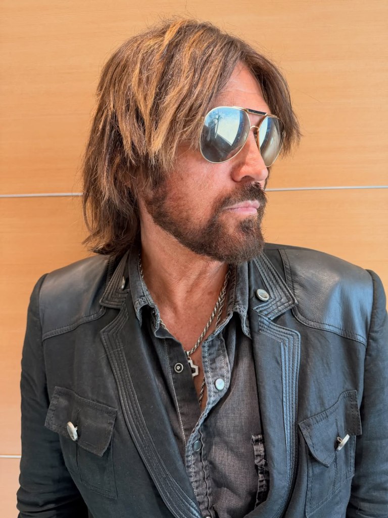 Billy Ray Cyrus wearing aviator sunglasses, a dark shirt, and a black leather jacket.