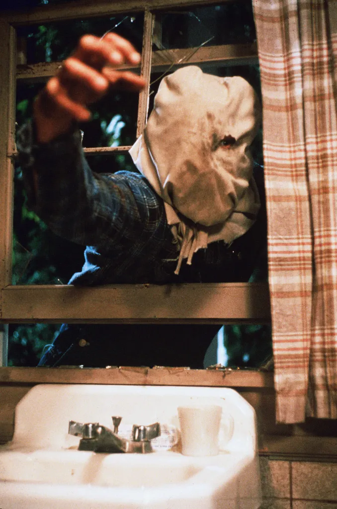 Warrington Gillette as Jason Voorhees wearing a sack over his head, reaching through a broken window above a bathroom sink in a scene from 