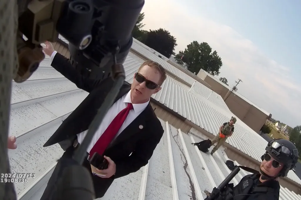 Bodycam footage shows law enforcement officers on a roof, with one in a suit and red tie speaking while gesturing.