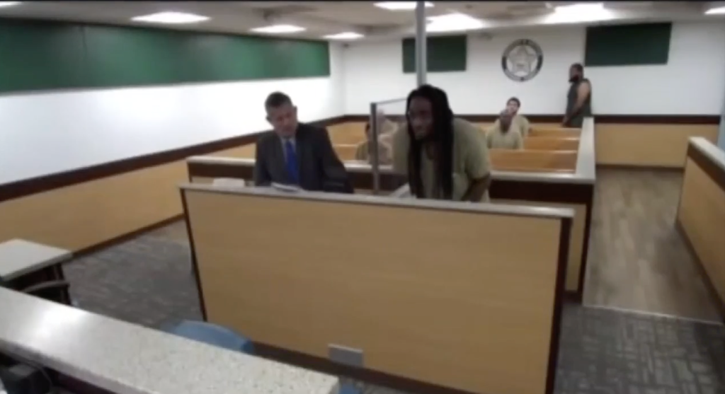 A man with dreadlocks in jail attire stands before a judge in a bond court hearing.