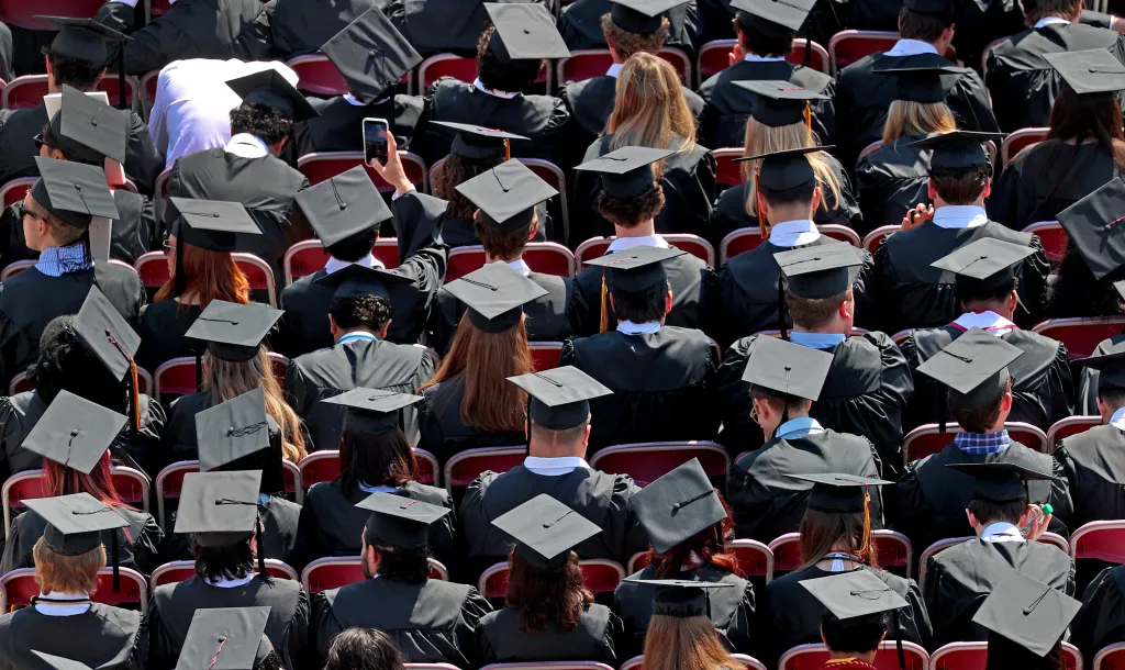 A high-angle view of graduating students wearing black gowns and mortarboards sitting in stadium seats.