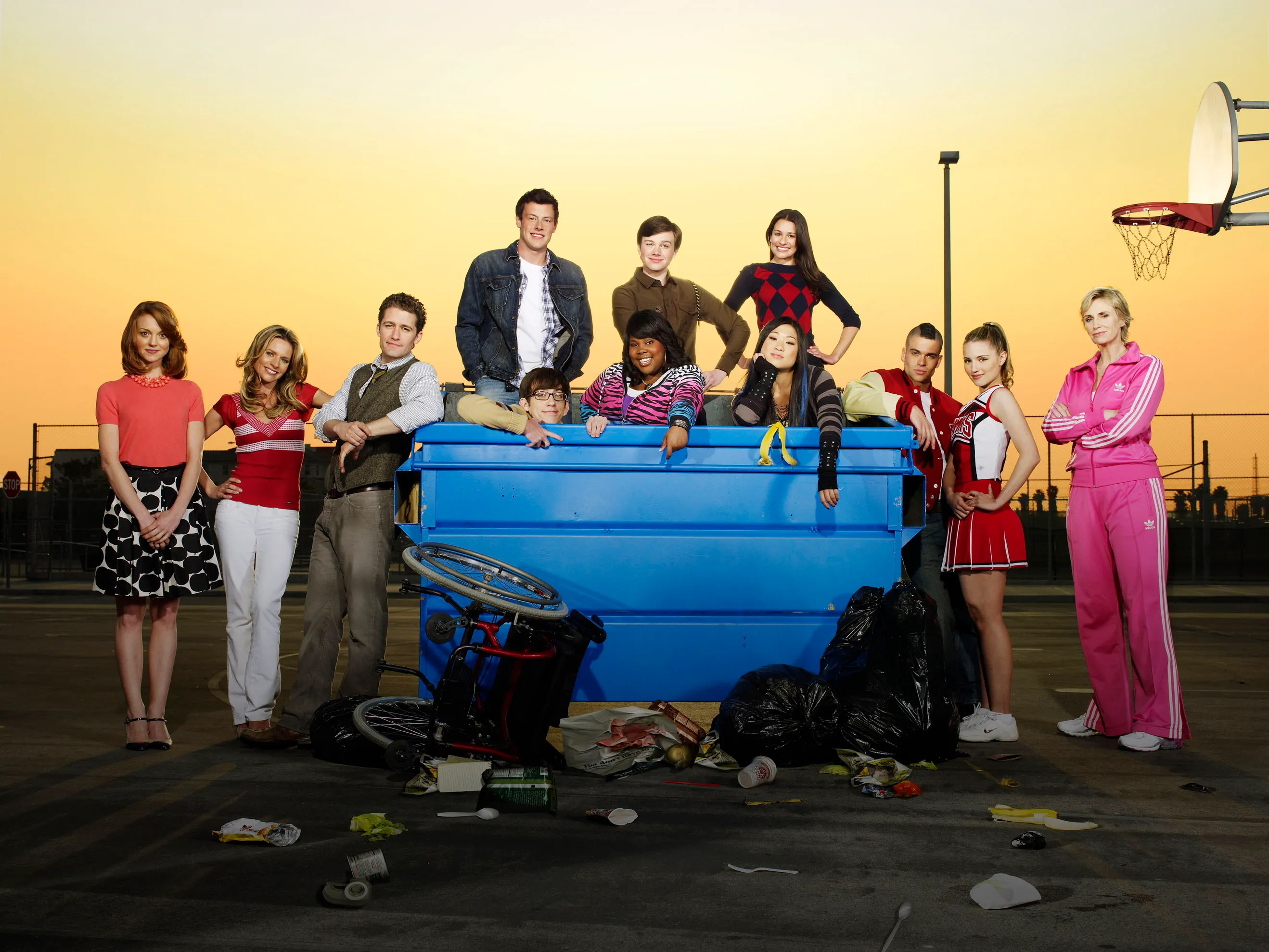Cast photo of the television show Glee, featuring the main characters posing around and inside a large blue dumpster outdoors at sunset.