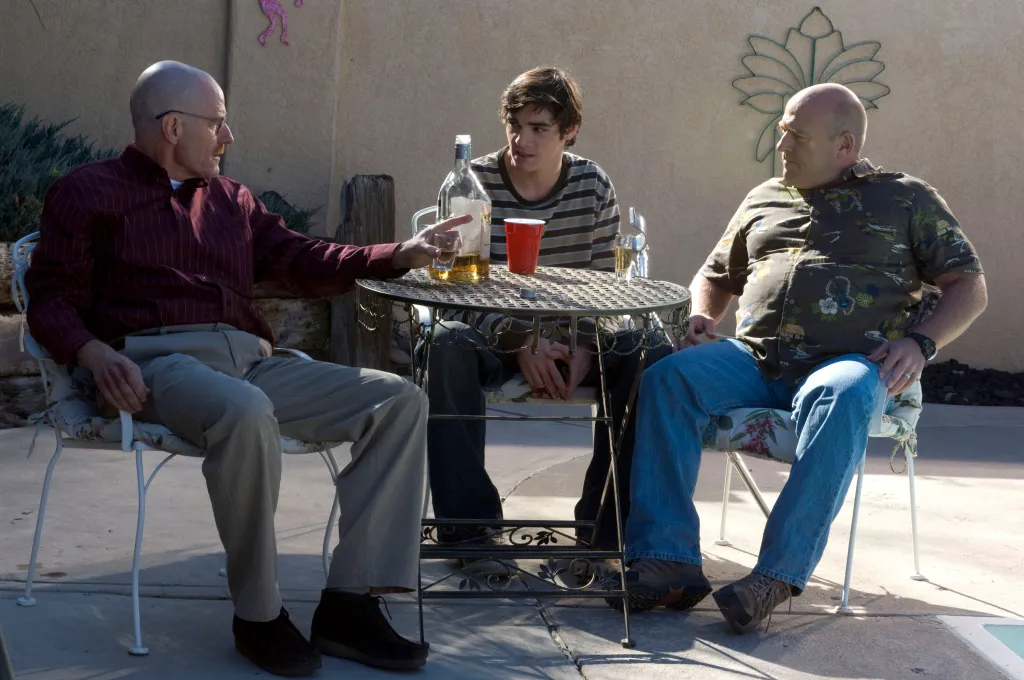 Bryan Cranston, RJ Mitte, and Dean Norris sitting at a table outdoors in a scene from Breaking Bad.