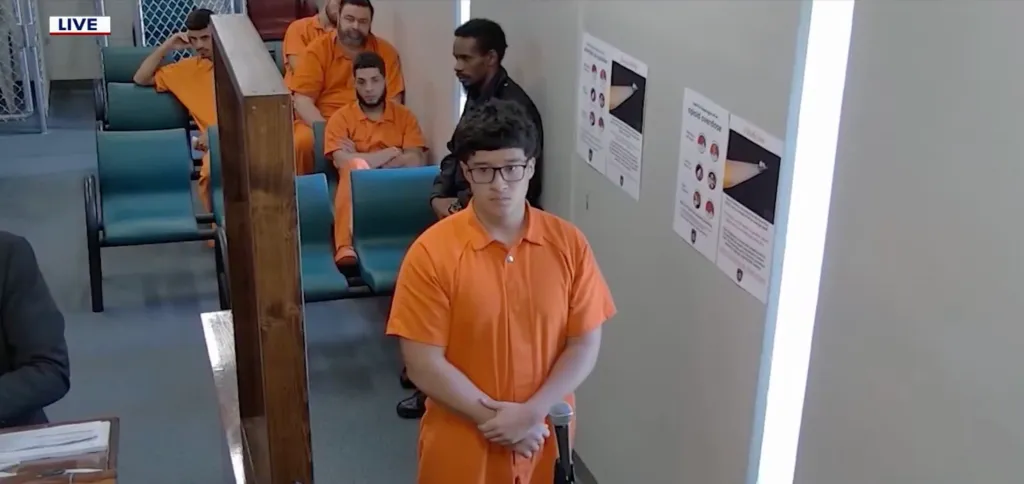 Brian Alexander Ramirez wearing an orange jumpsuit and glasses, standing before a judge with other defendants seated behind him.