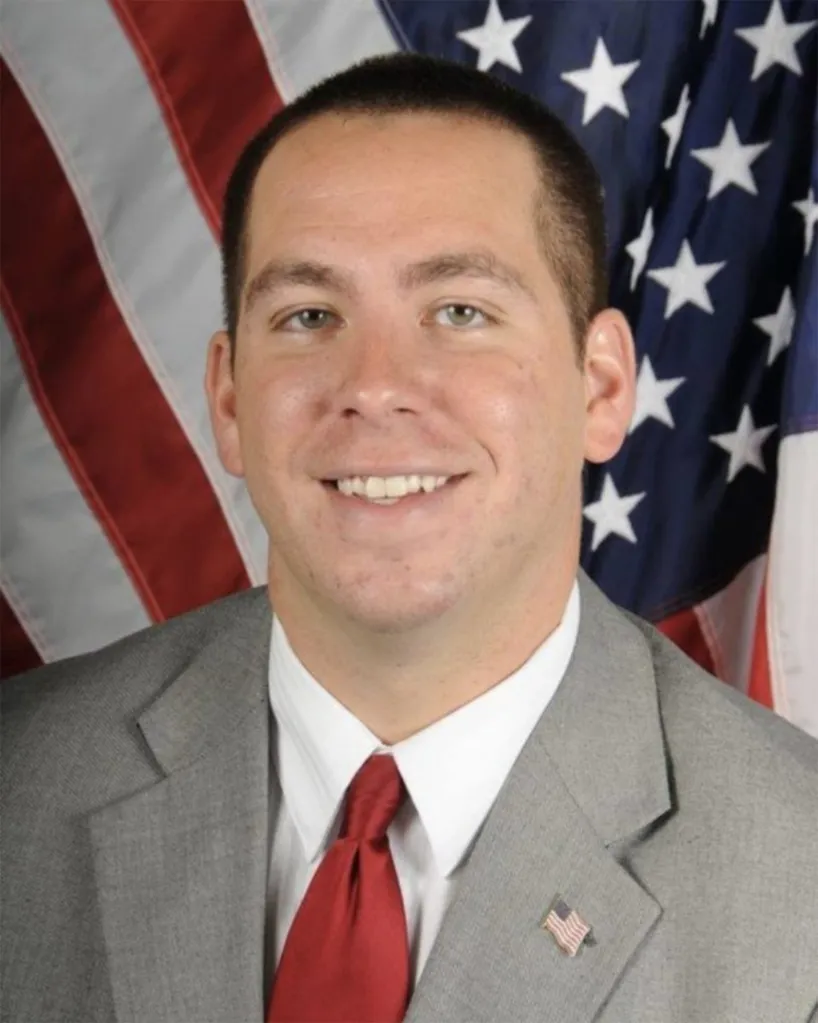 Portrait of Brian Maher in a grey suit and red tie, with an American flag in the background.