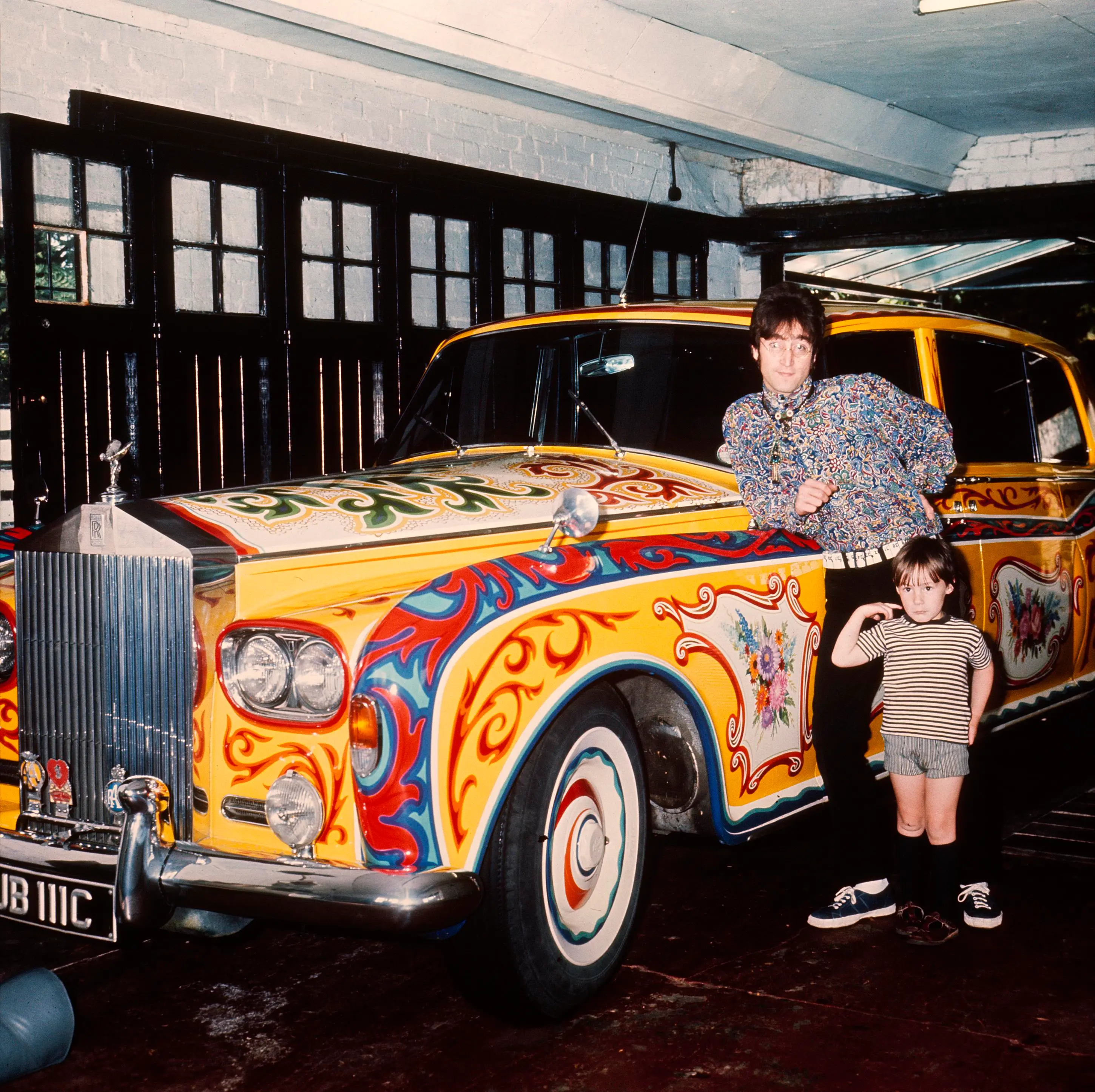 John Lennon and his son Julian posing next to their psychedelic painted Rolls-Royce in a garage.