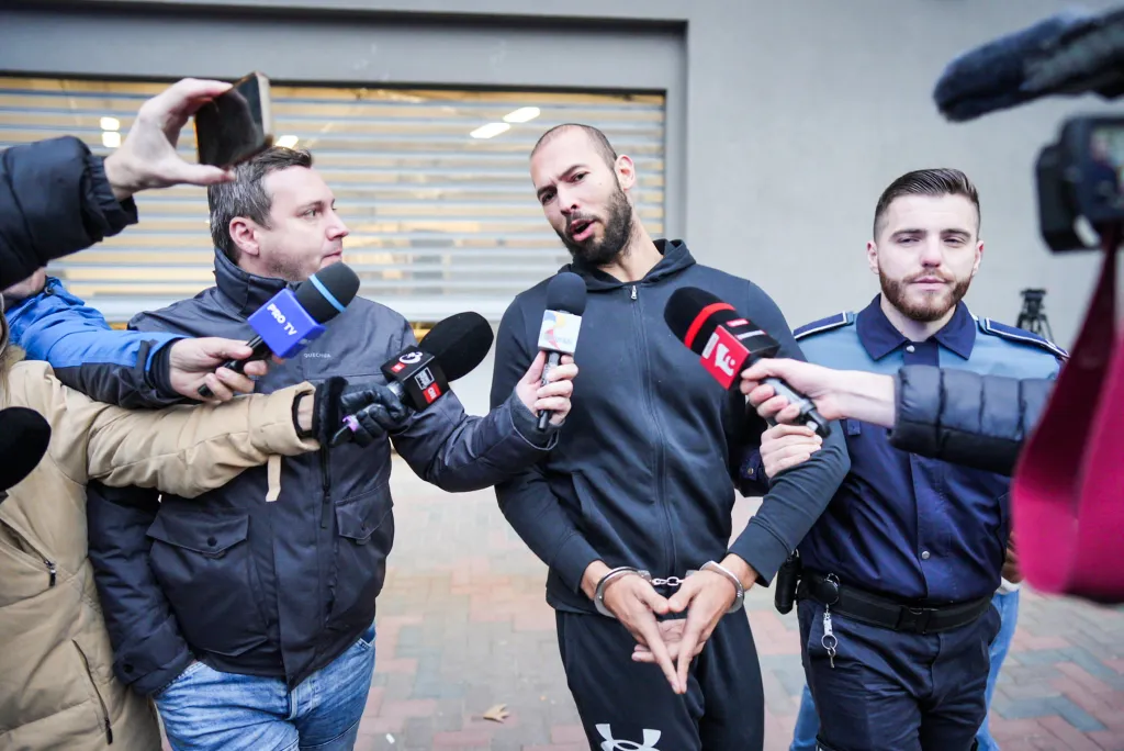 Andrew Tate talks to media while surrounded by reporters and a police officer.