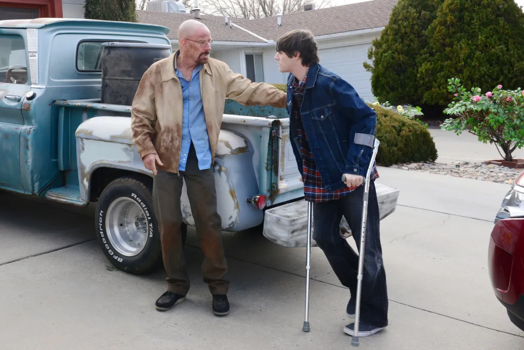 Bryan Cranston as Walter White putting a hand on RJ Mitte's shoulder as Walter Jr. stands with crutches next to a blue pickup truck.