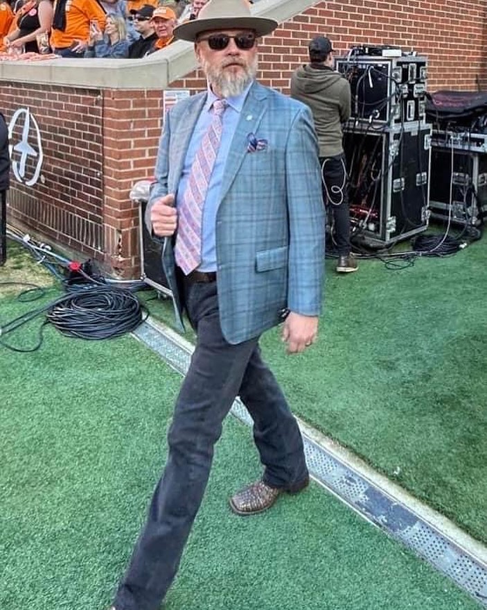 A man with a beard, hat, and sunglasses walks across artificial turf, wearing a plaid sport coat, light blue shirt, tie, and jeans.