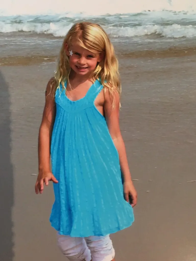 A young blonde girl in a blue dress standing on a beach.
