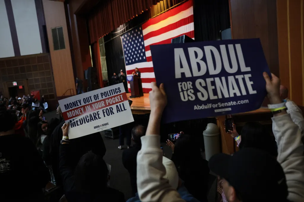Supporters hold campaign signs for Senate candidate Abdul El-Sayed at a rally in front of an American flag.
