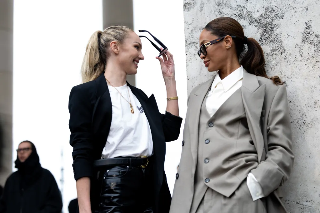 Candice Swanepoel and Joan Smalls stand outside during Paris Fashion Week, wearing stylish outfits and sunglasses.
