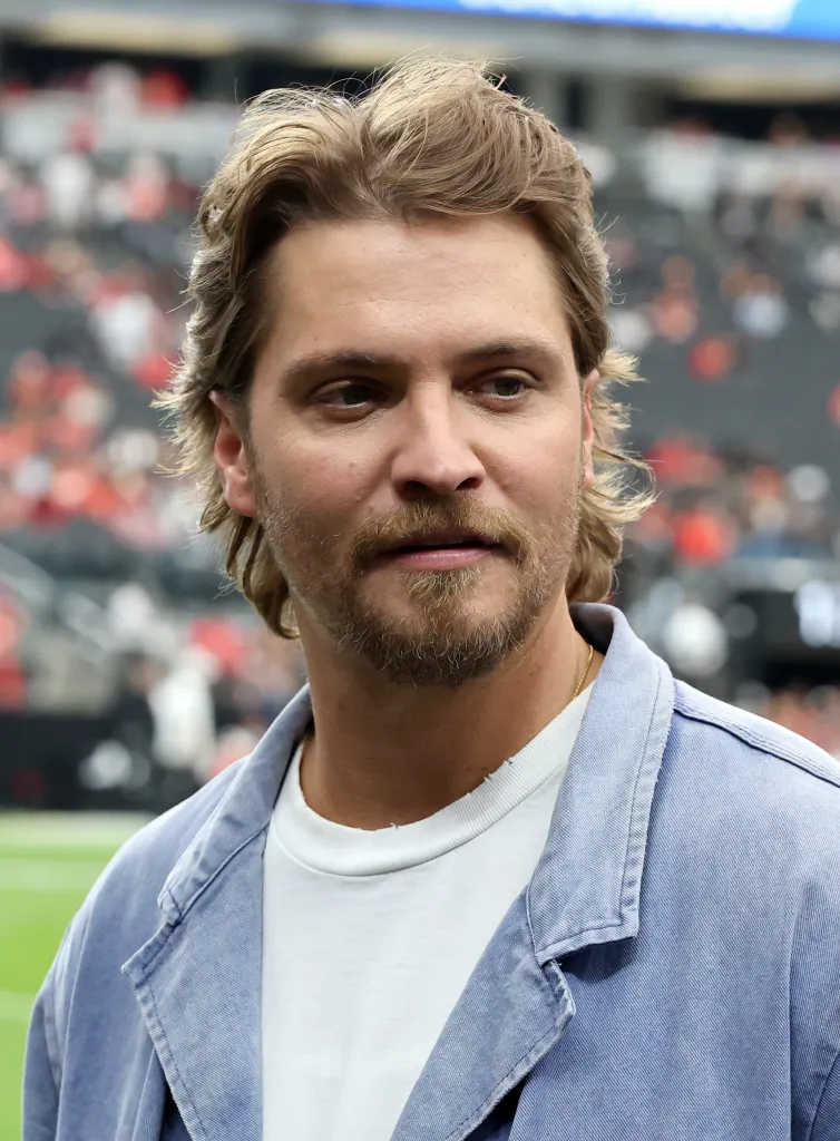 Cast member Luke Grimes stands on a sideline at a football stadium.