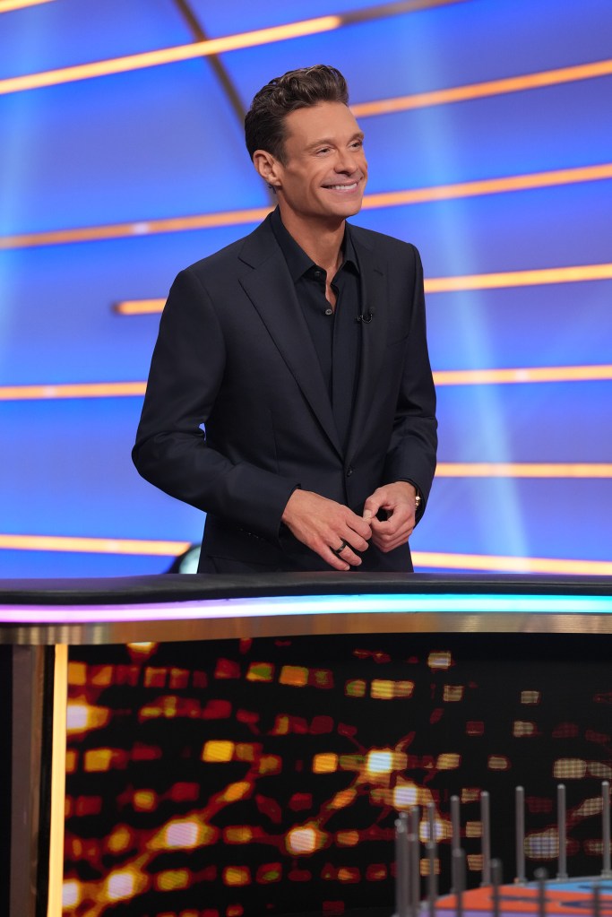 Ryan Seacrest stands smiling behind a brightly lit game show set on Celebrity Wheel of Fortune.