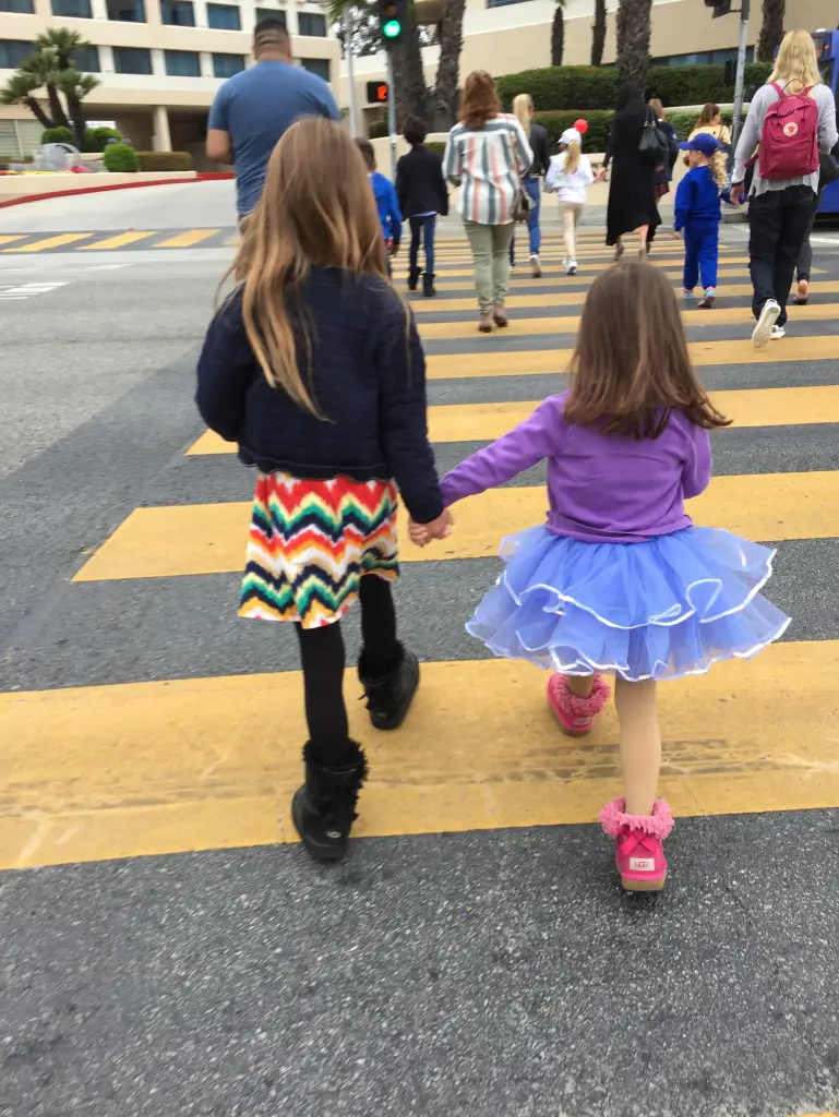 Two young girls hold hands while walking across a crosswalk.