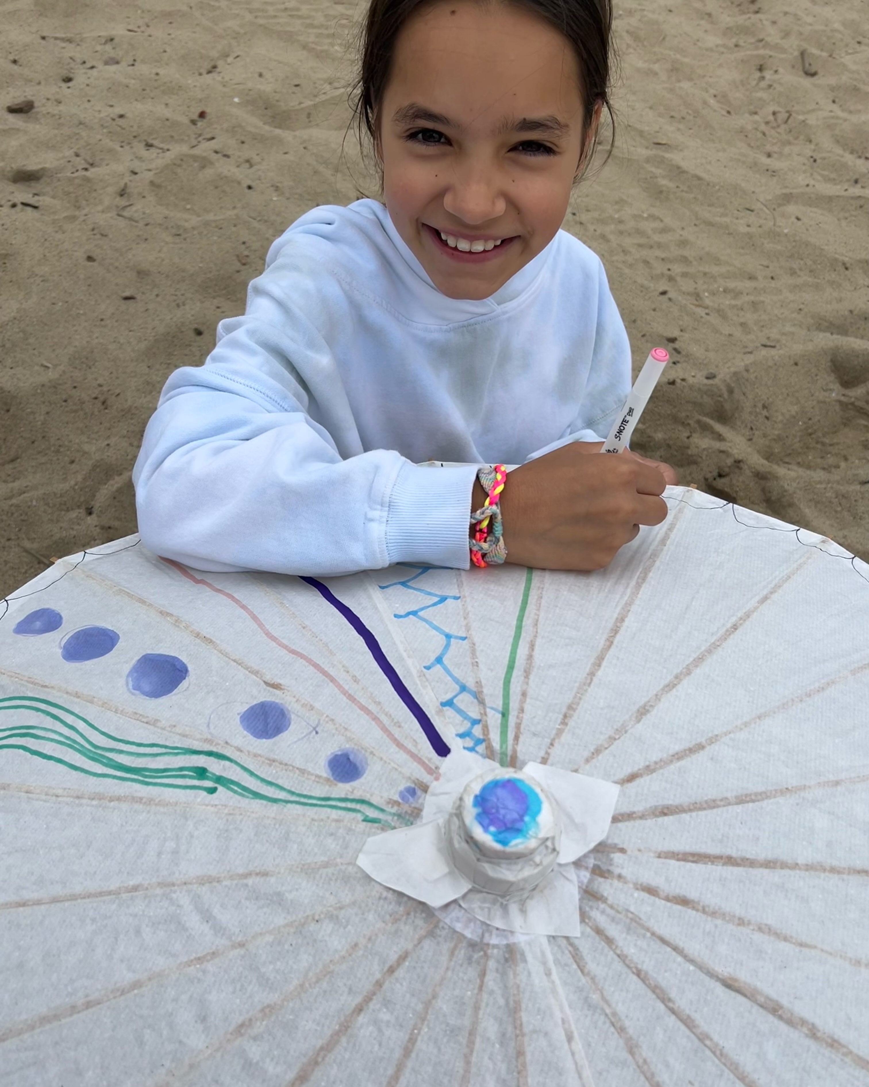 A young girl smiles as she sits in the sand, drawing on a white umbrella with a marker.