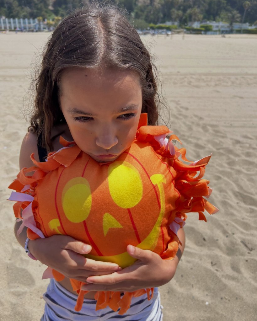 A young girl at the beach holding a plush, orange pumpkin pillow with a sad facial expression.