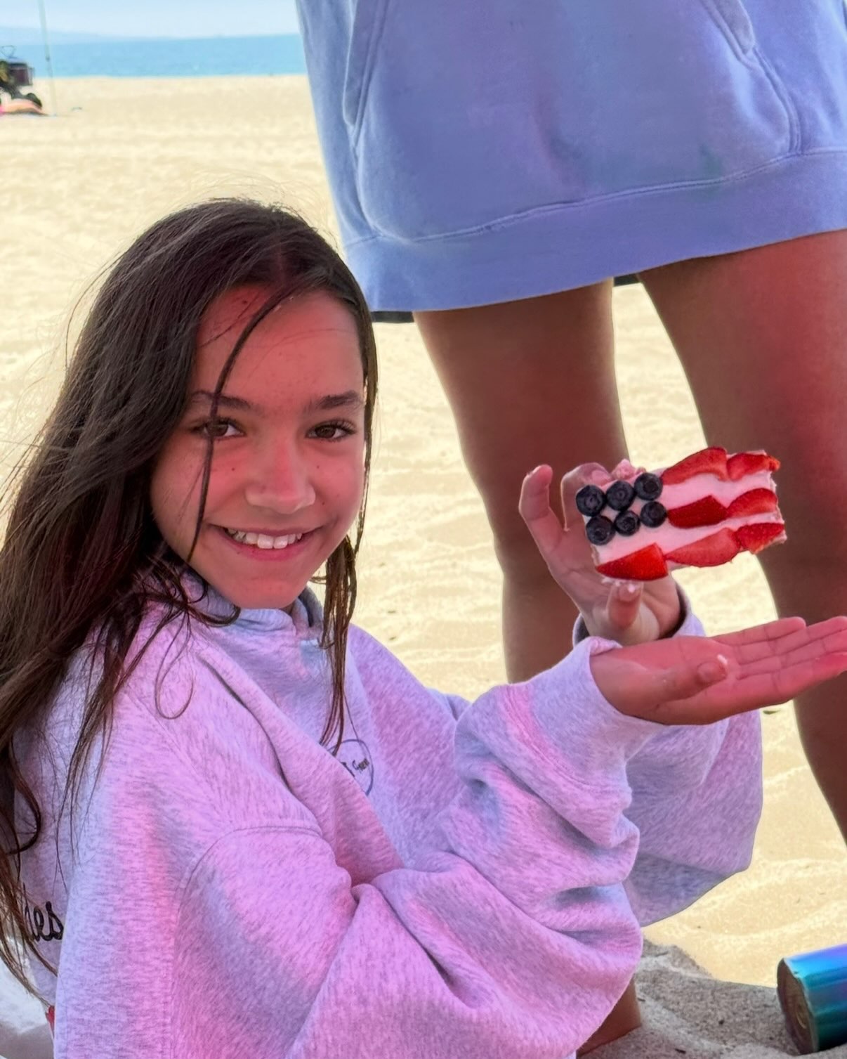 Fiona Lowe smiling while holding a small cake decorated with an American flag made of strawberries and blueberries at the beach.